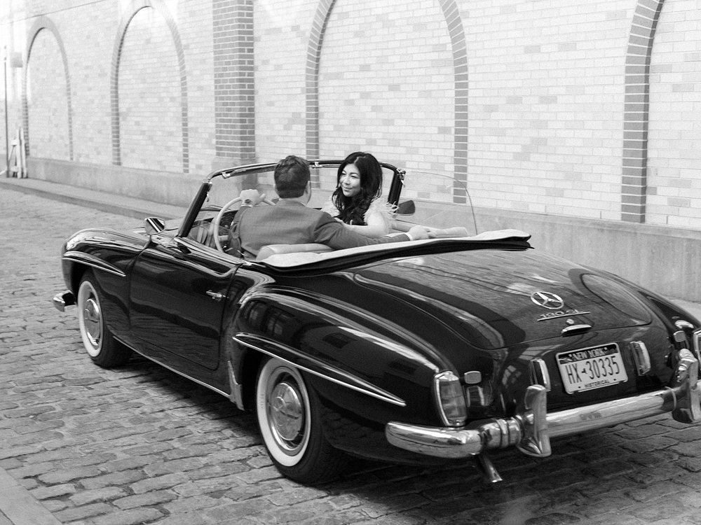 woman and man sit in front of classic Mercedes Benz 190sl while woman looks back over shoulder 