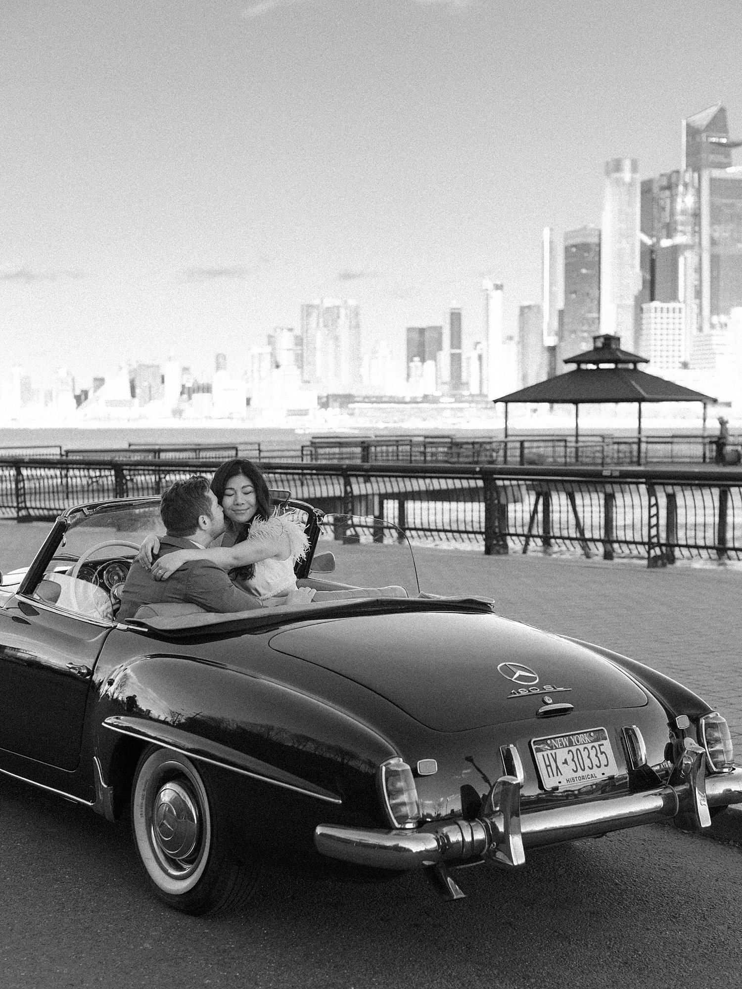man holds fiancee on lap kissing her cheek at Maxwell Park with Manhattan skyline behind them inside classic Mercedes Benz 190sl