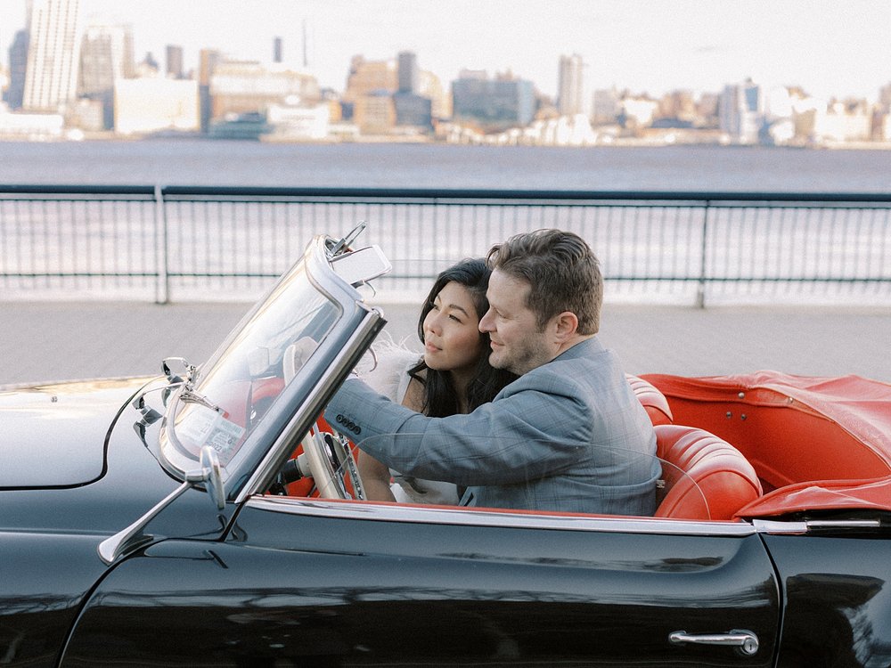 man drives fiancee along Maxwell Park with Manhattan skyline behind them