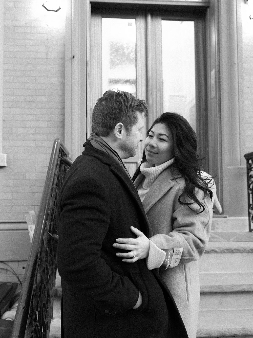 engaged couple stands on steps of Hoboken NJ apartment bundled in jackets