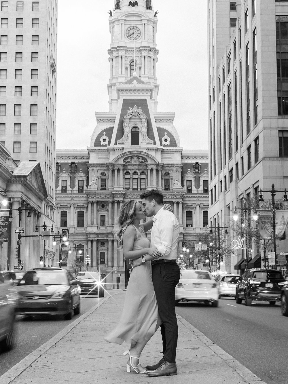 engaged couple stands in front of Philadelphia City Hall at night with traffic around them
