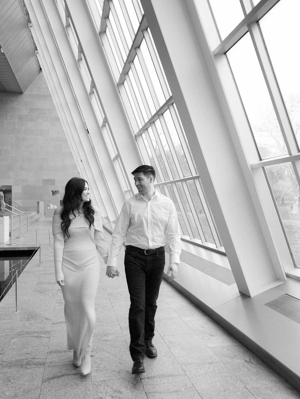 engaged couple holds hands walking by windows in the Temple of Dendur exhibit at the Metropolitan Museum of Art