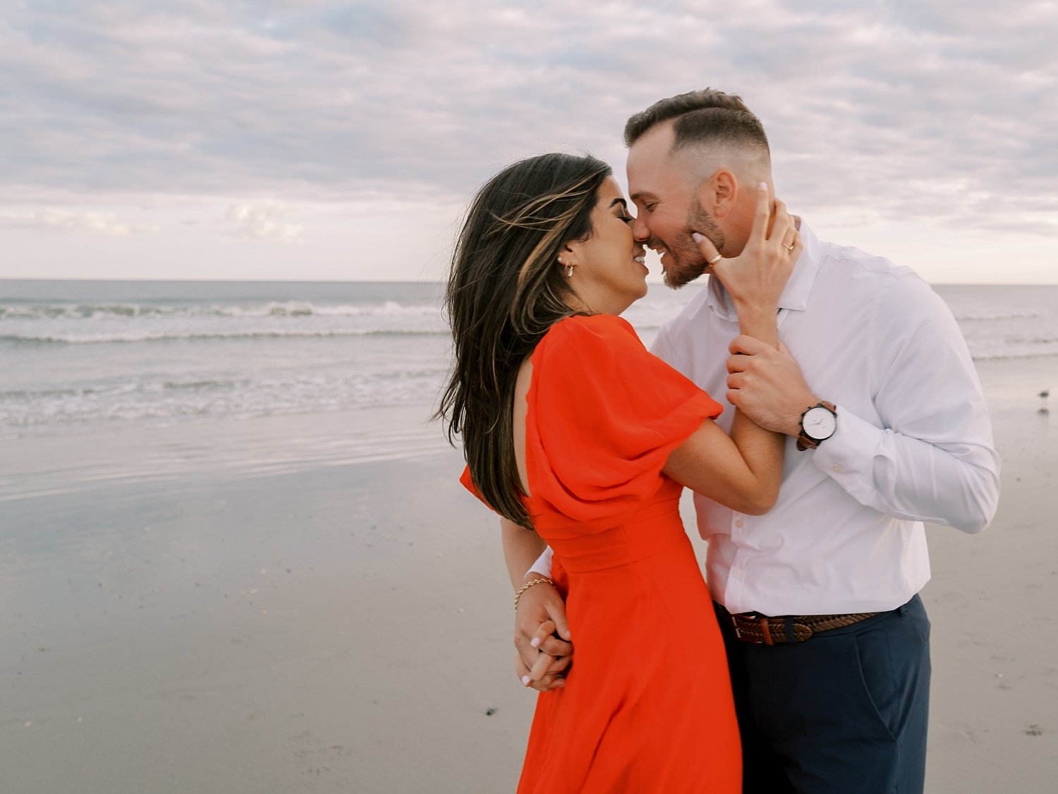 man in white shirt leans to kiss woman in orange dress during Ocean City NJ engagement session on beach