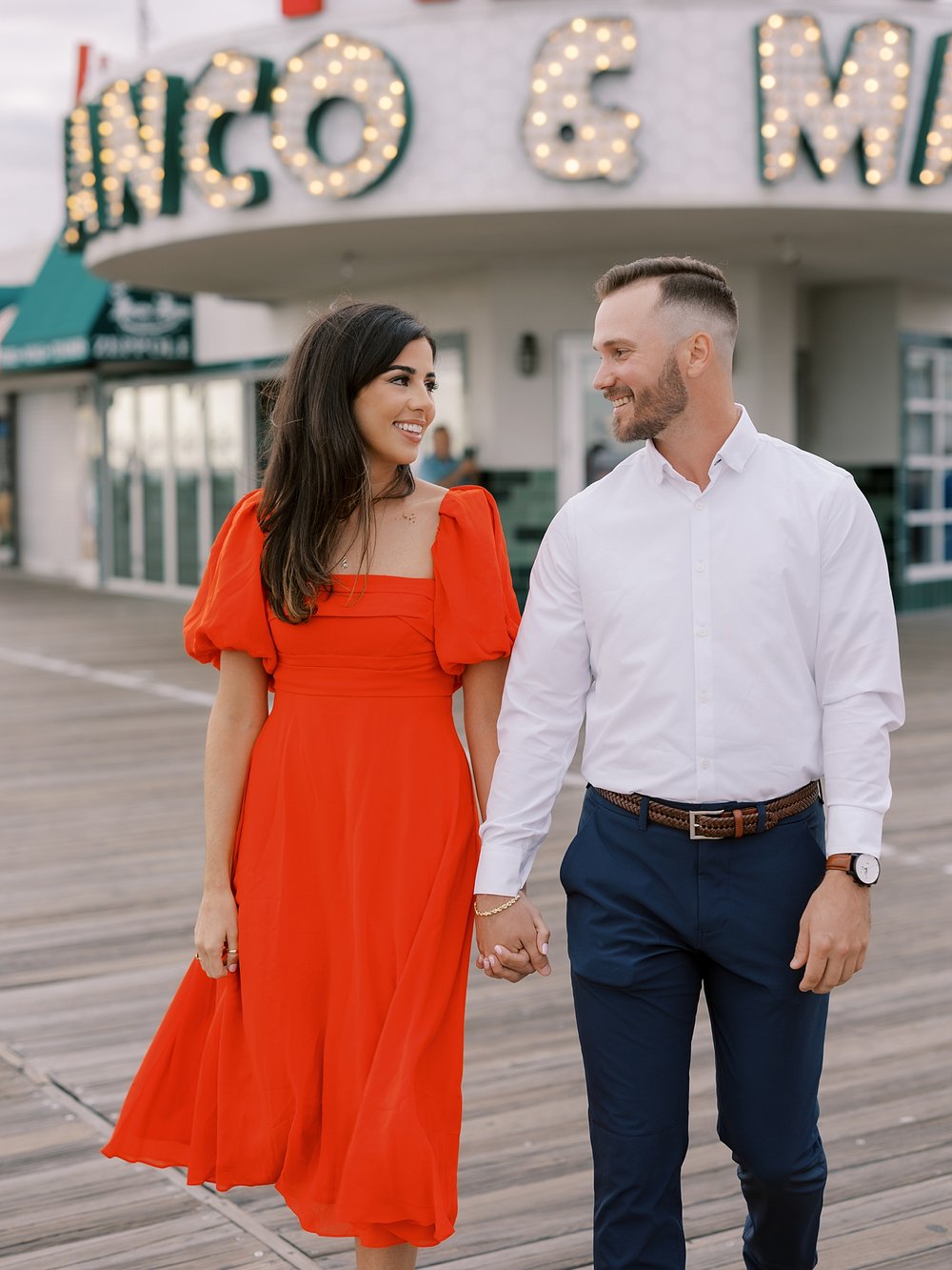 engaged couple holds hands walking in front of lit signs on Ocean City Boardwalk