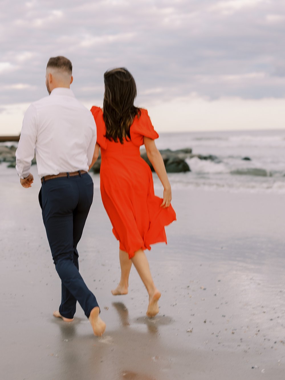 woman in orange dress holds skirt walking on beach with fiancee in Ocean City NJ