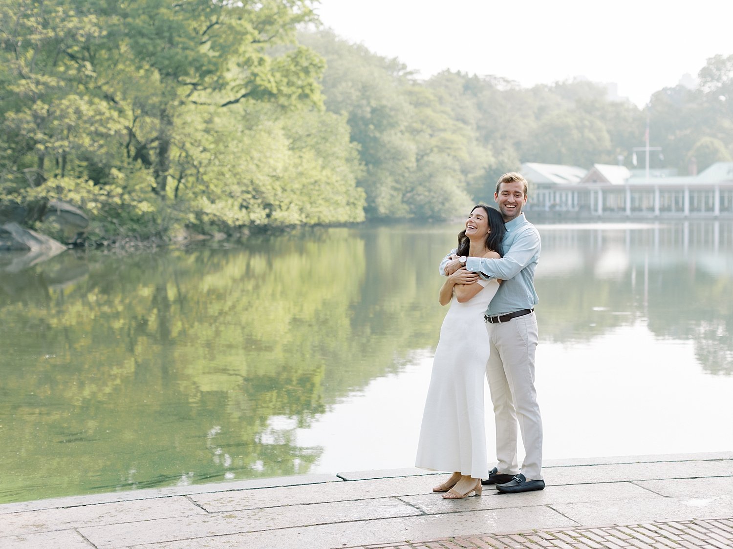 engaged couple hugs in front of Central Park boathouse by lake