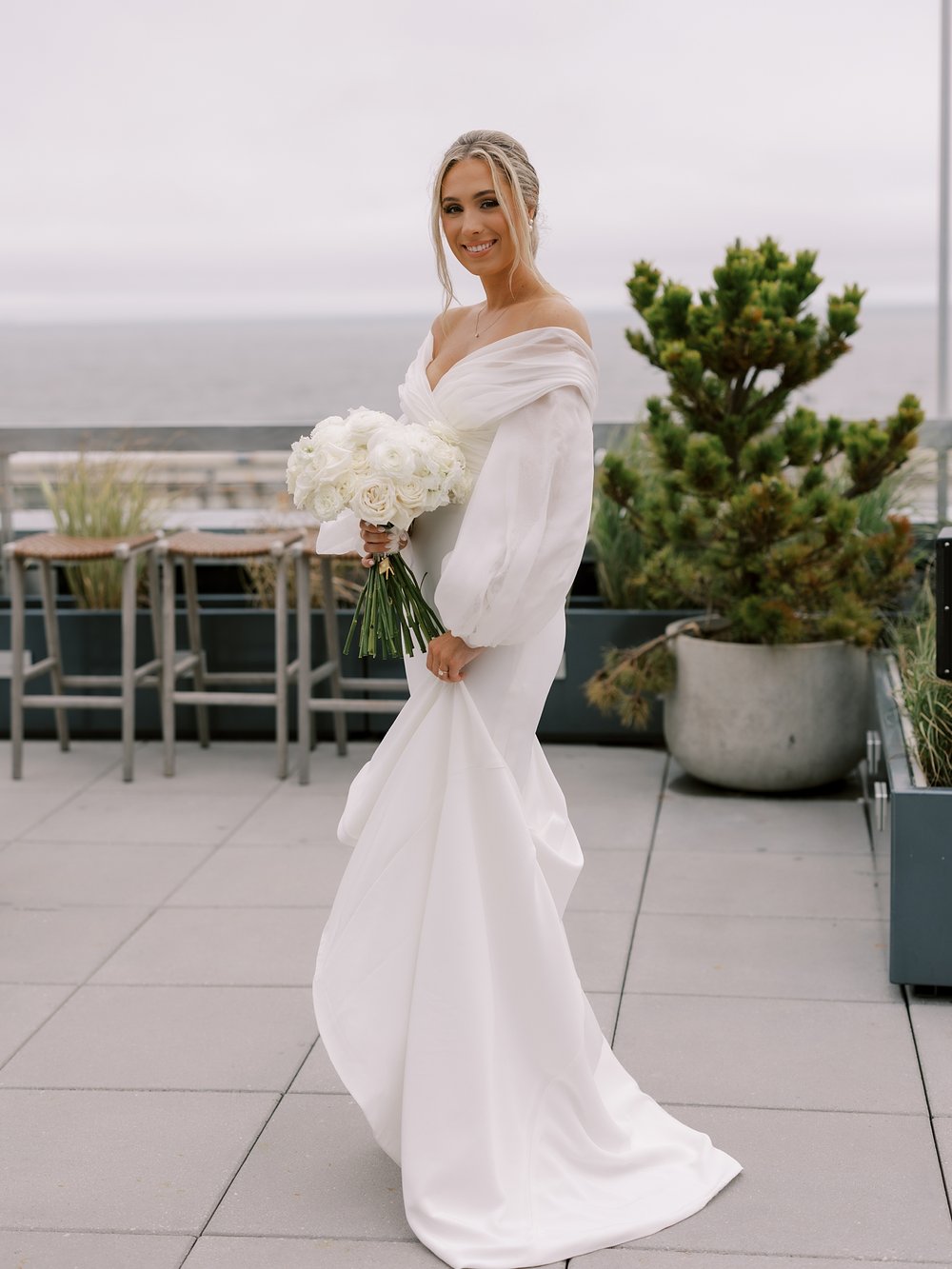 bride stands on patio at the Rockaway Hotel with off-the-shoulder wedding gown and bouquet of ivory flowers 