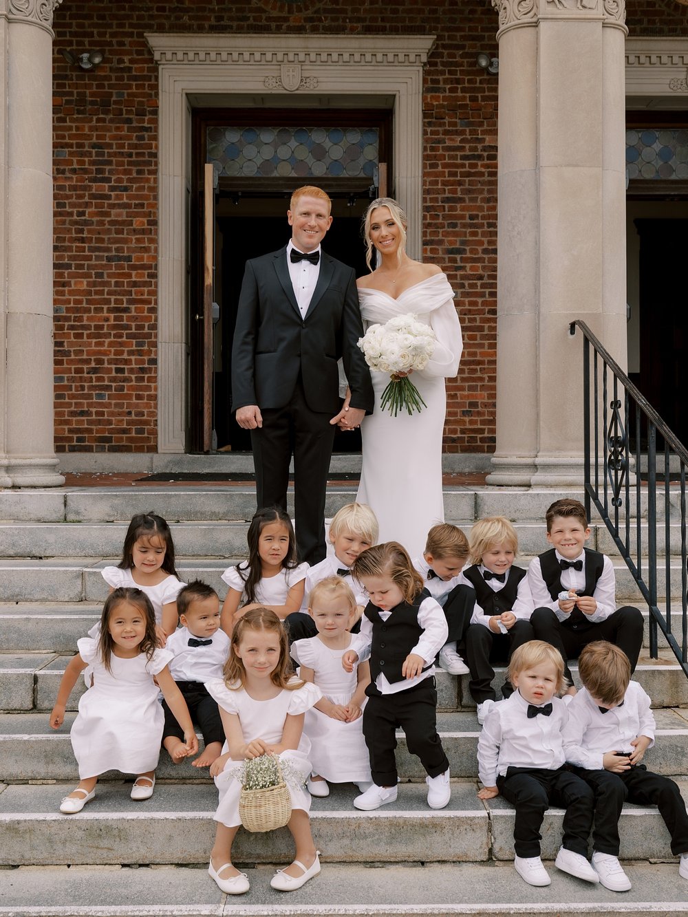 bride and groom sit on steps with flower girls and ring bearers outside Long Island NY church