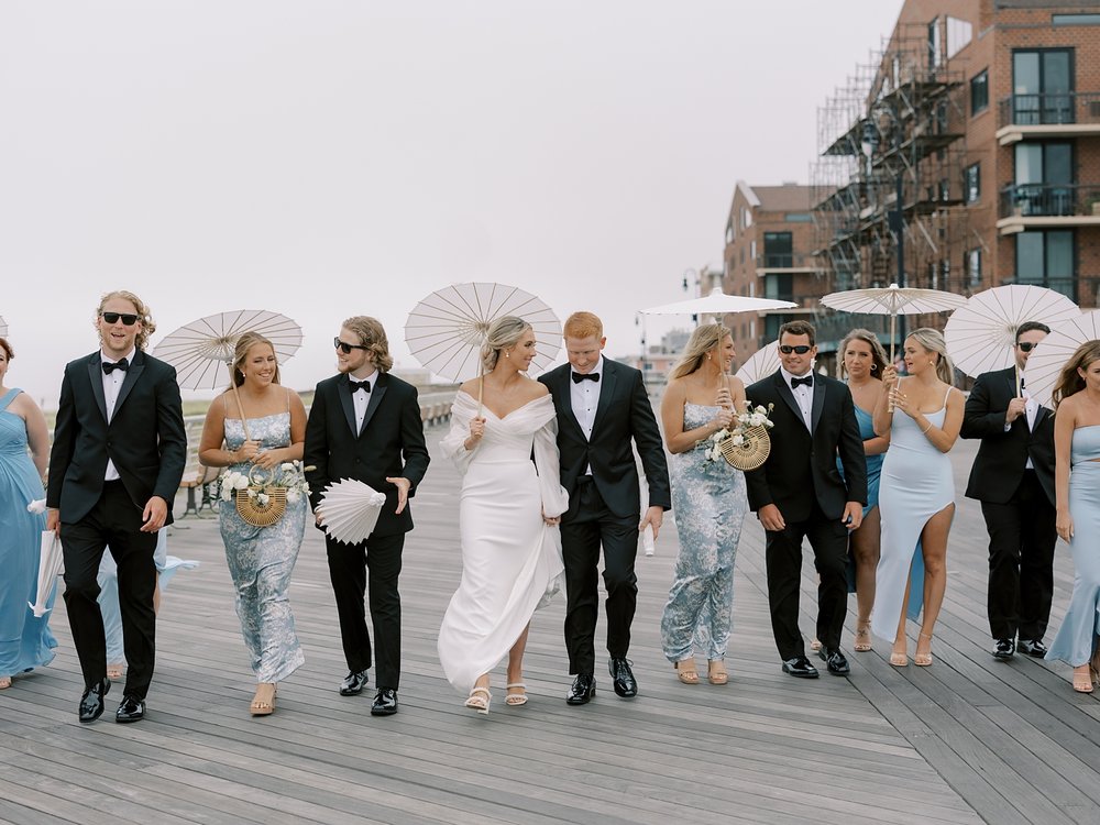 bride and groom walk with wedding party in black suits and blue and white dresses on Long Island boardwalk