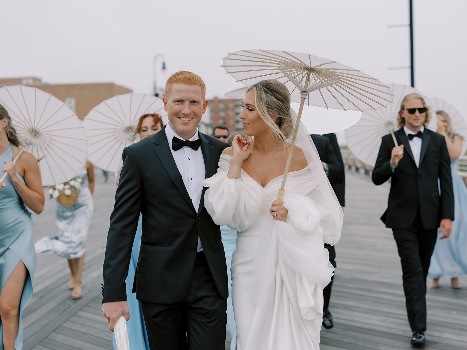 bride and groom walk on Long Island beach boardwalk with wedding party carrying white parasols 