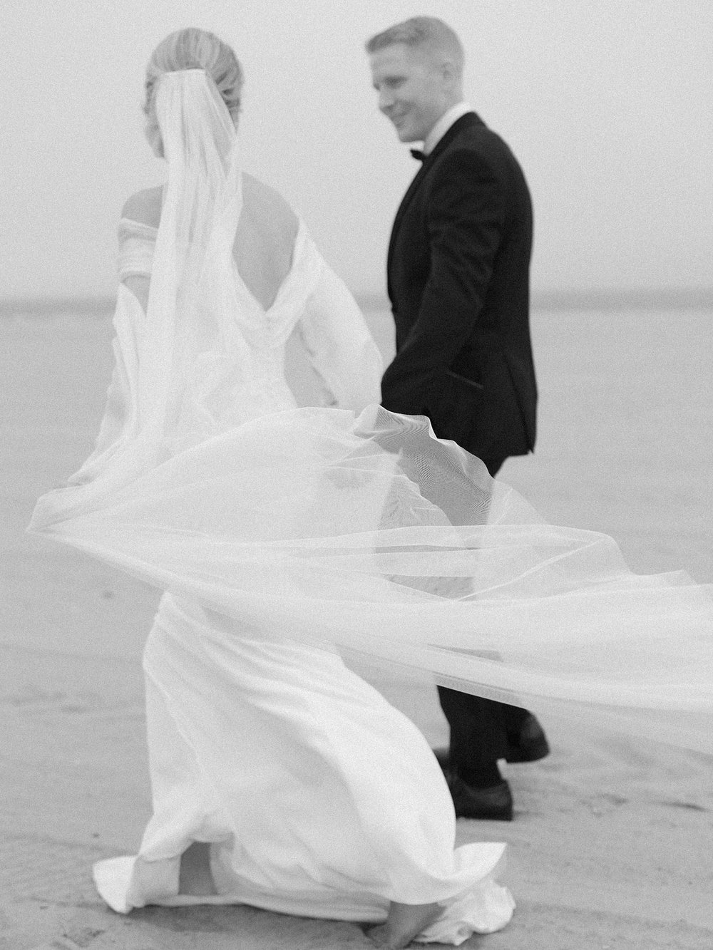 bride and groom hold hands walking on beach with veil floating around them on Long Island beach