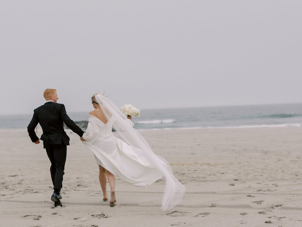 bride and groom hold hands walking on windy day on Long Island beach