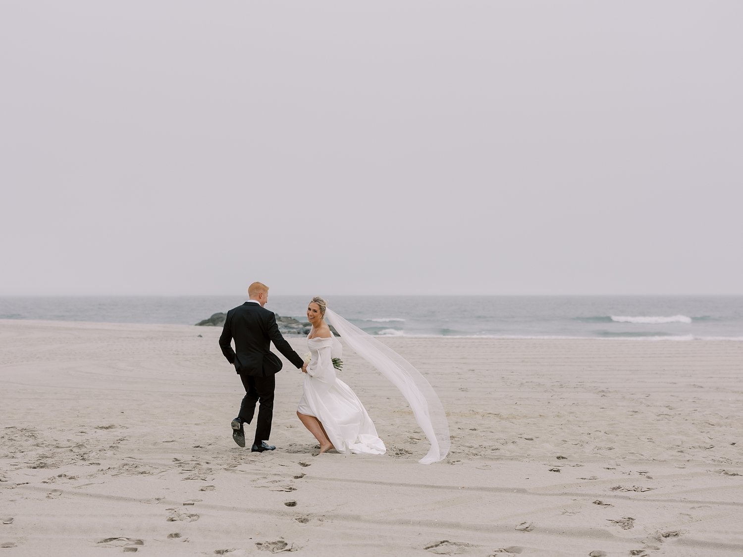 bride and groom run on Long Island beach holding hands on windy day