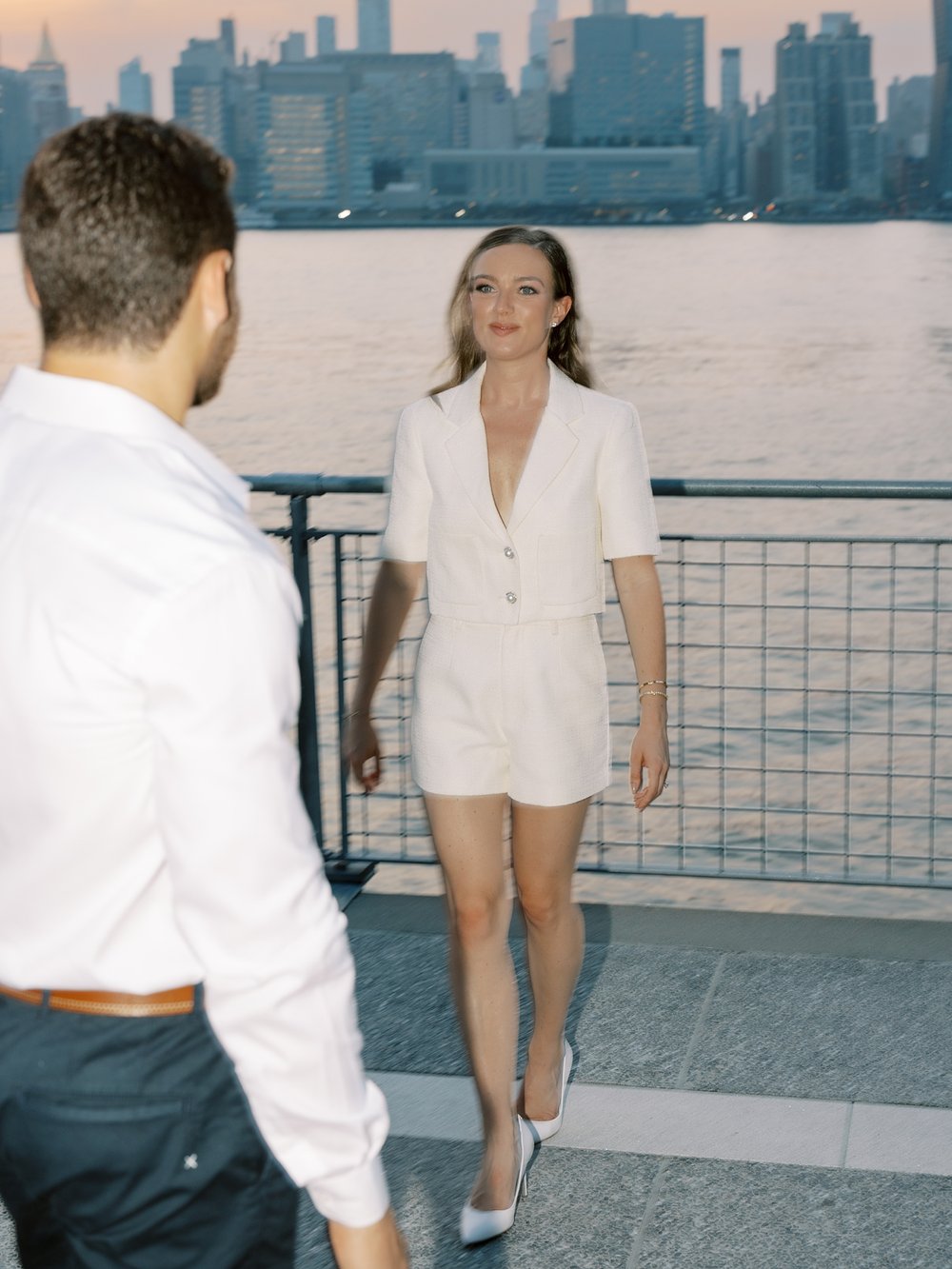 woman in ivory Revolve pantsuit walks to fiancee inside Greenpoint Waterfront Park