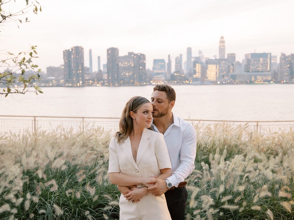 man hugs woman from behind in front of fence at Greenpoint Waterfront Park