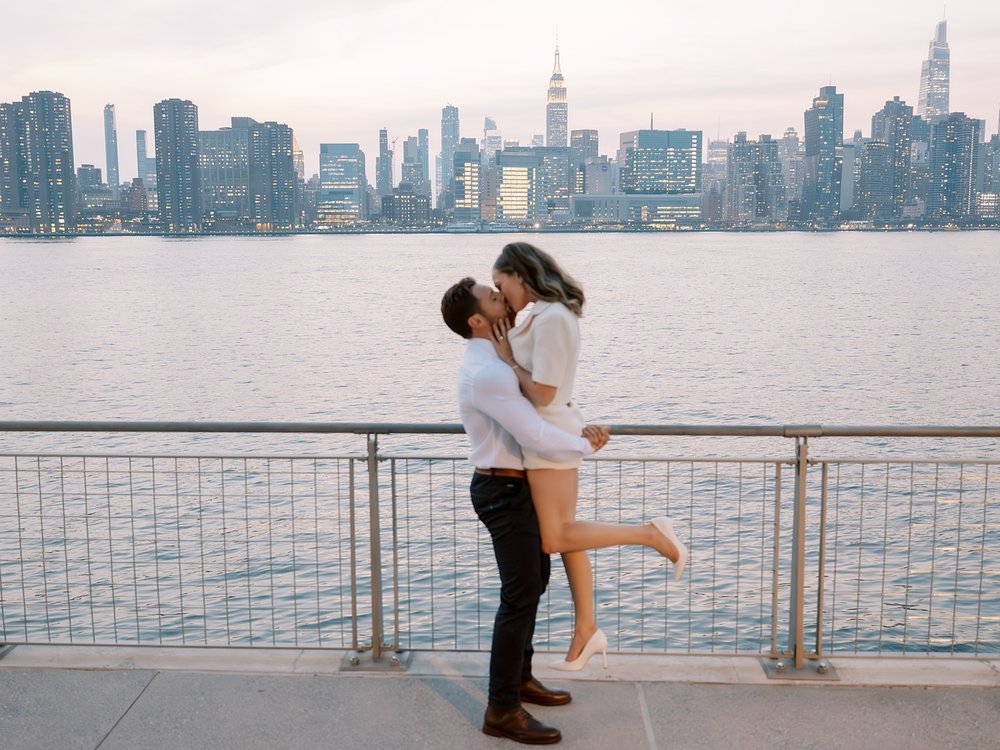man lifts up fiancee in front of railing in Greenpoint Waterfront Park
