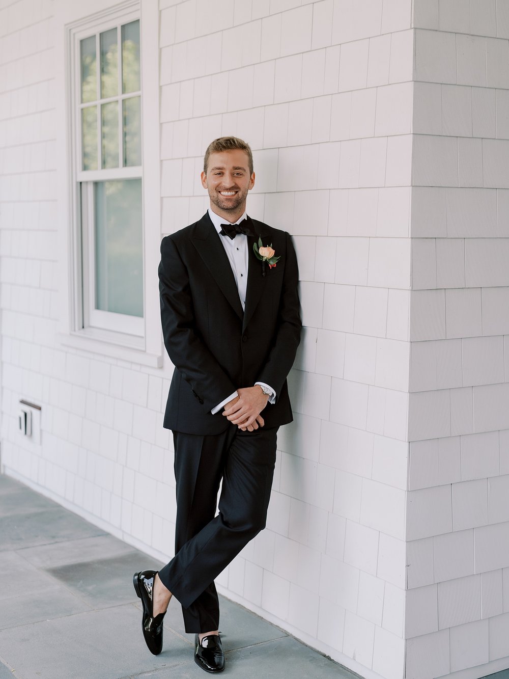 groom in black suit leans against white clapboard of The Country Club of New Canaan