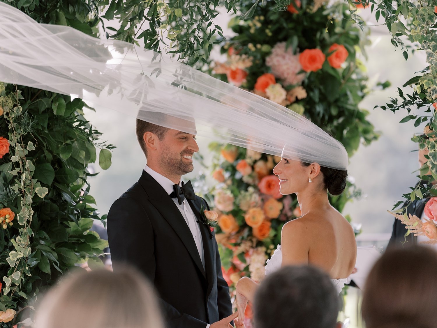 bride and groom hold hands in front of bright floral arbor with veil floating around them 