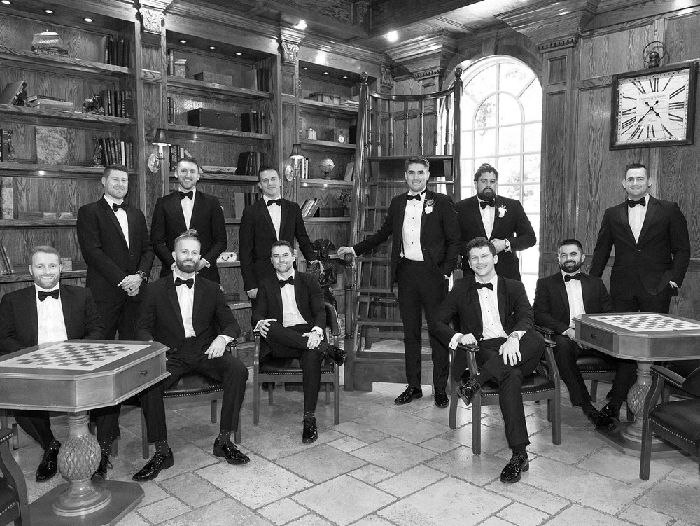 groom and groomsmen hold whiskey glasses sitting in library at the Ashford Estate