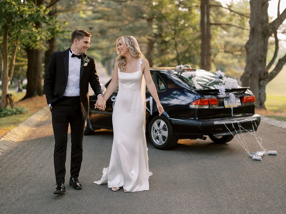bride and groom hold hands walking in front of groom's black car with "just married" sign