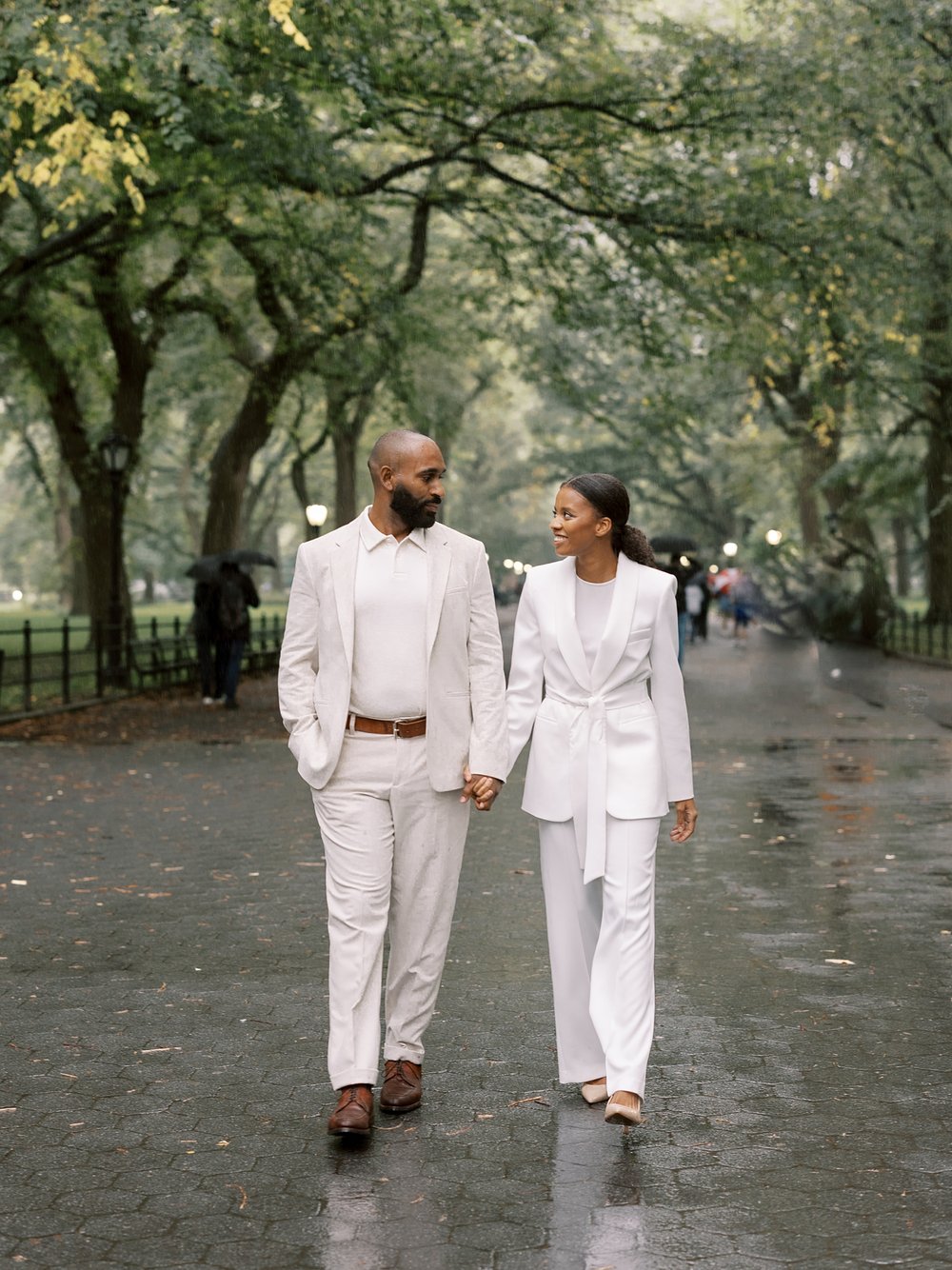 married couple in white suits hold hands walking through Central Park
