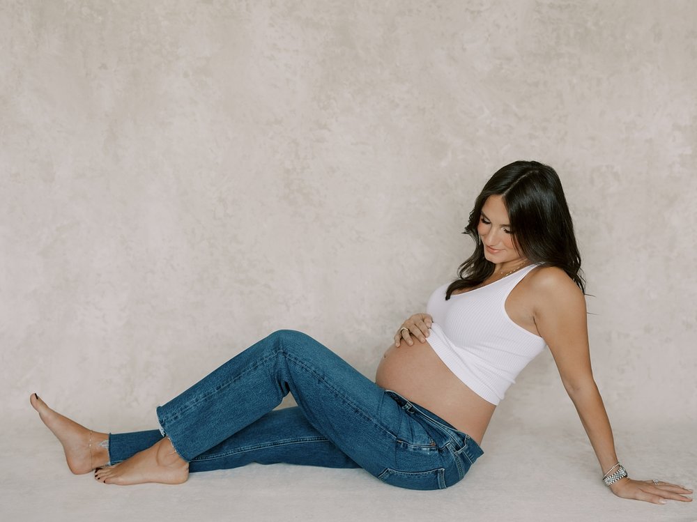 expecting mom sits holding baby bump in jeans and white bra during maternity photos