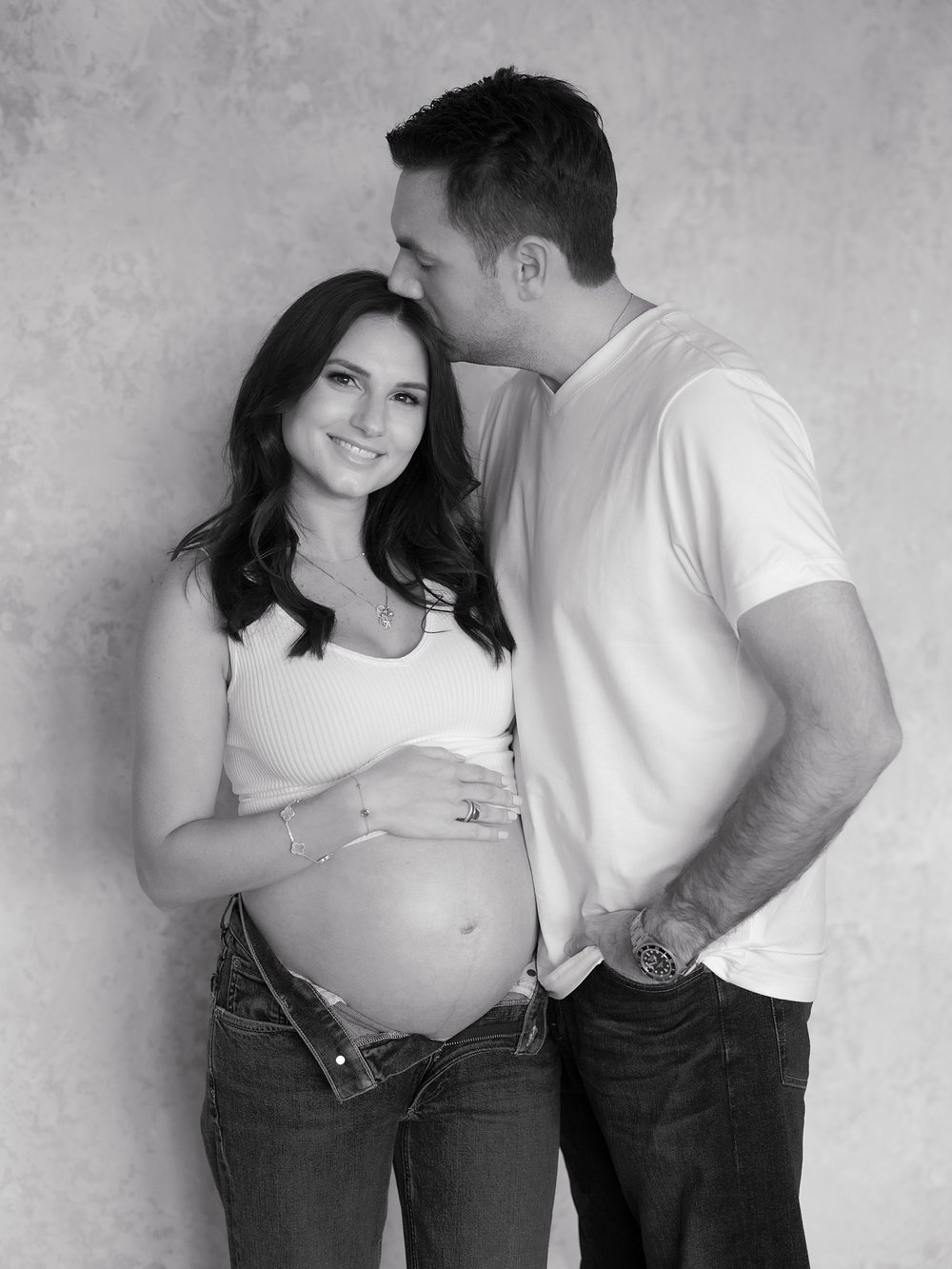 husband kisses wife's forehead while she holds baby bump during studio maternity session