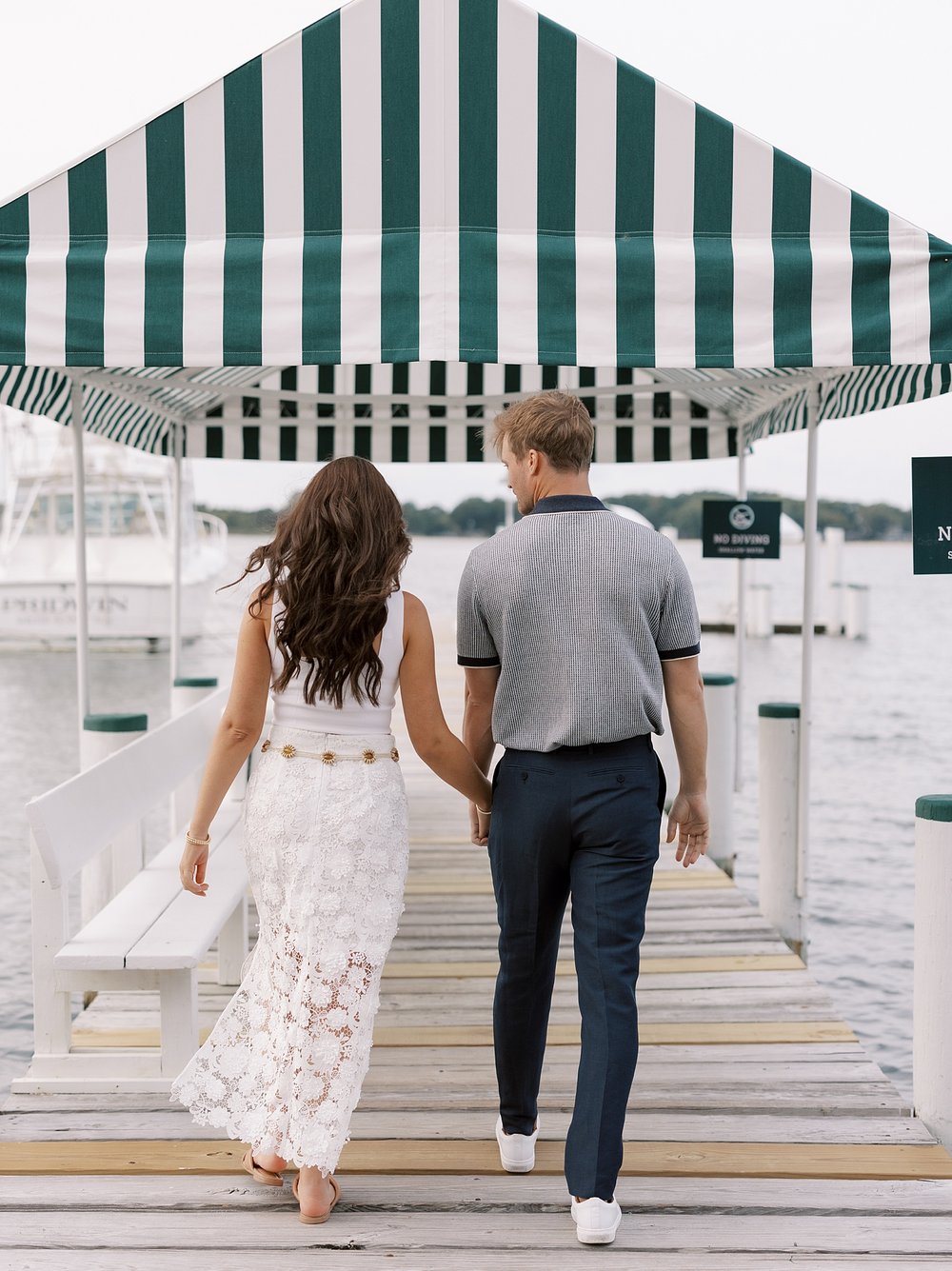 couple holds hands on dock walking under green and white awning