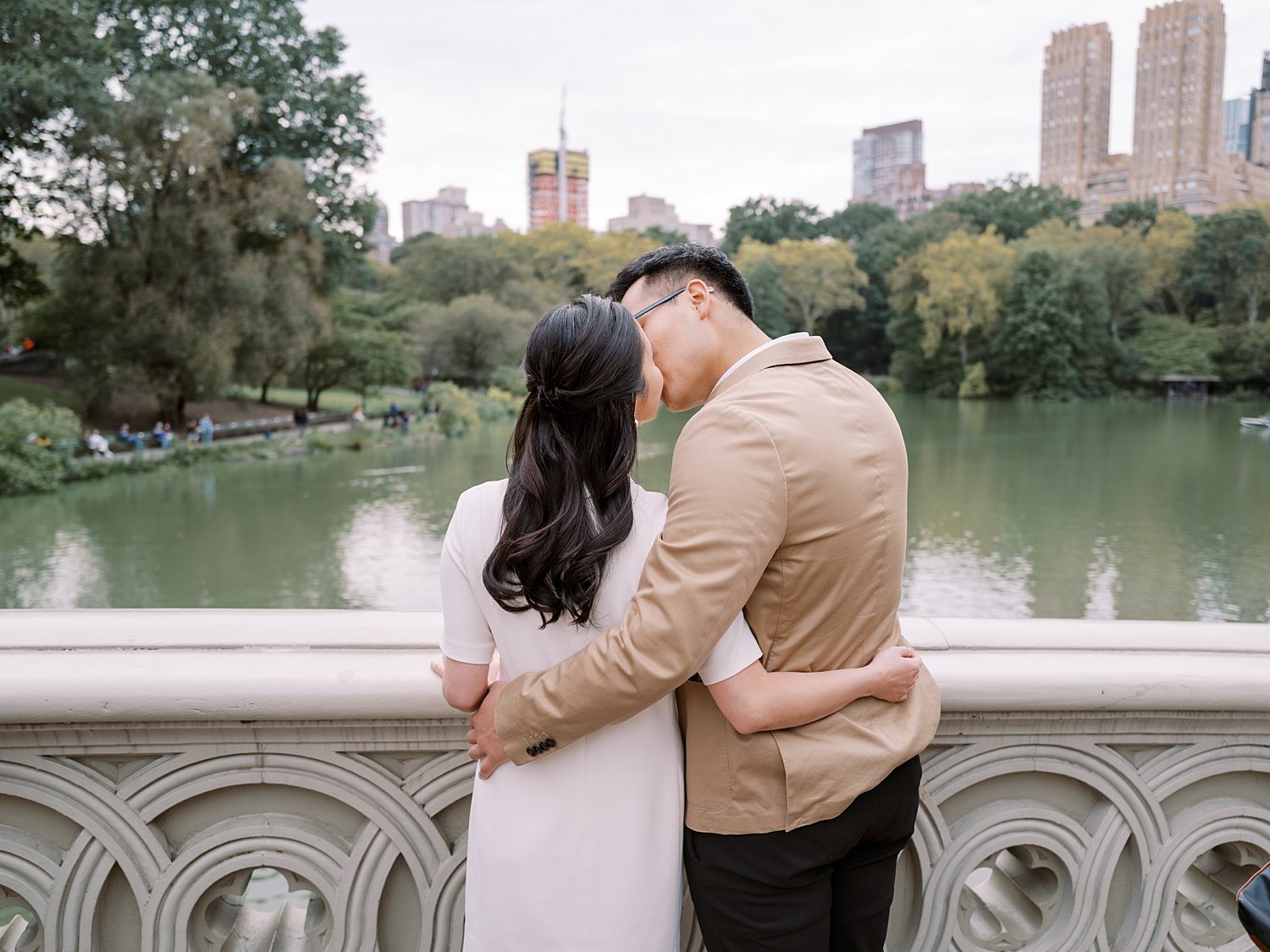 couple kisses in front of railing inside Central Park in front of NYC skyline