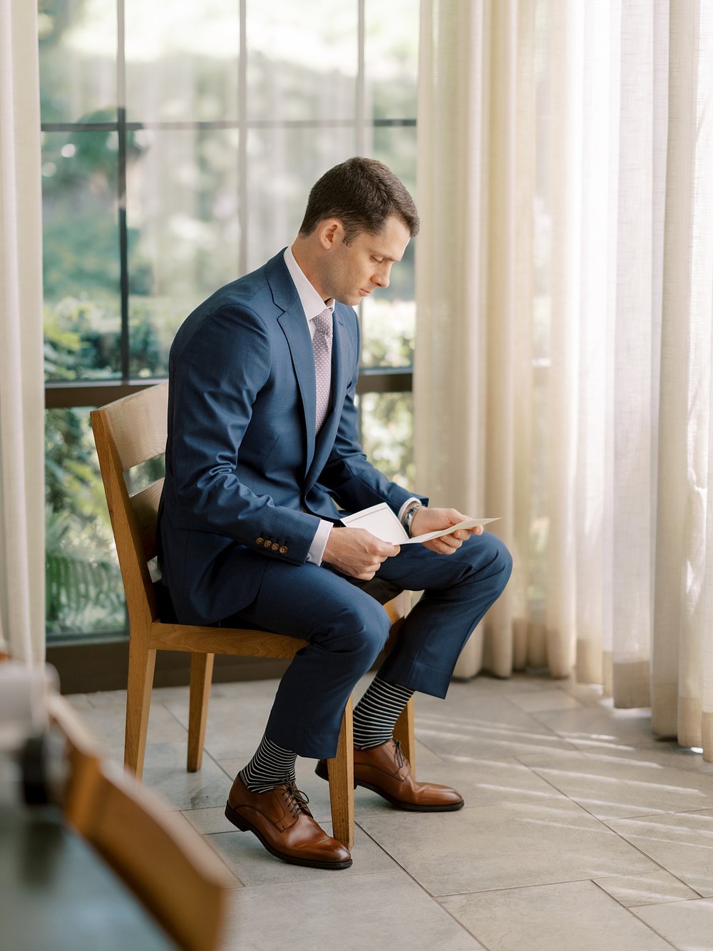 groom in navy suit reads letter sitting in chair by window
