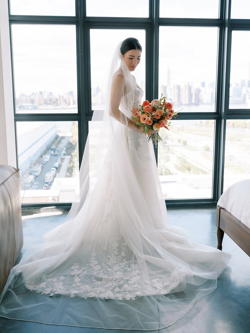 bride in strapless gown stands by window overlooking Brooklyn 