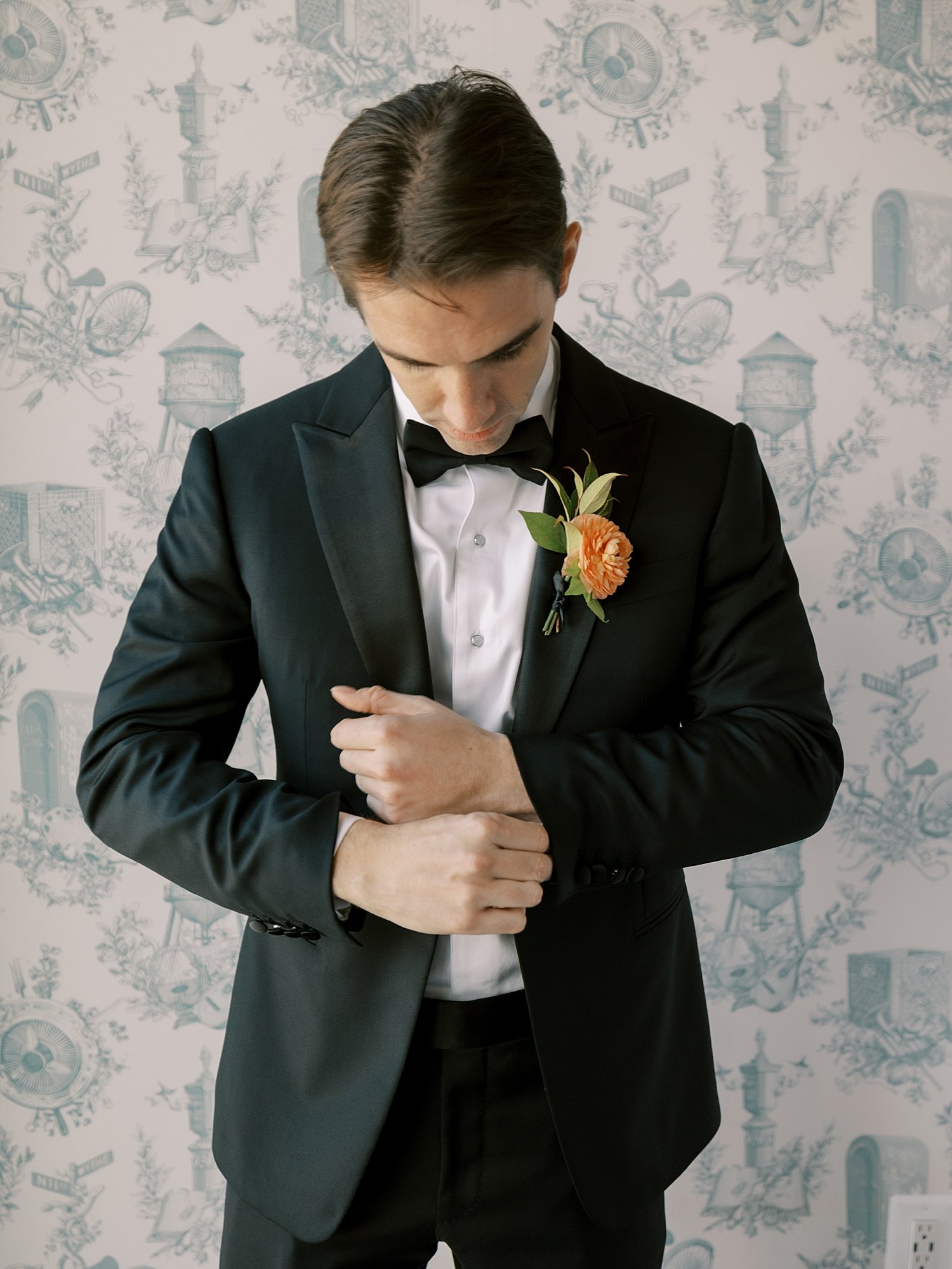 groom adjusts cufflinks in front of blue and white wallpaper at the Wythe Hotel in Brooklyn NY