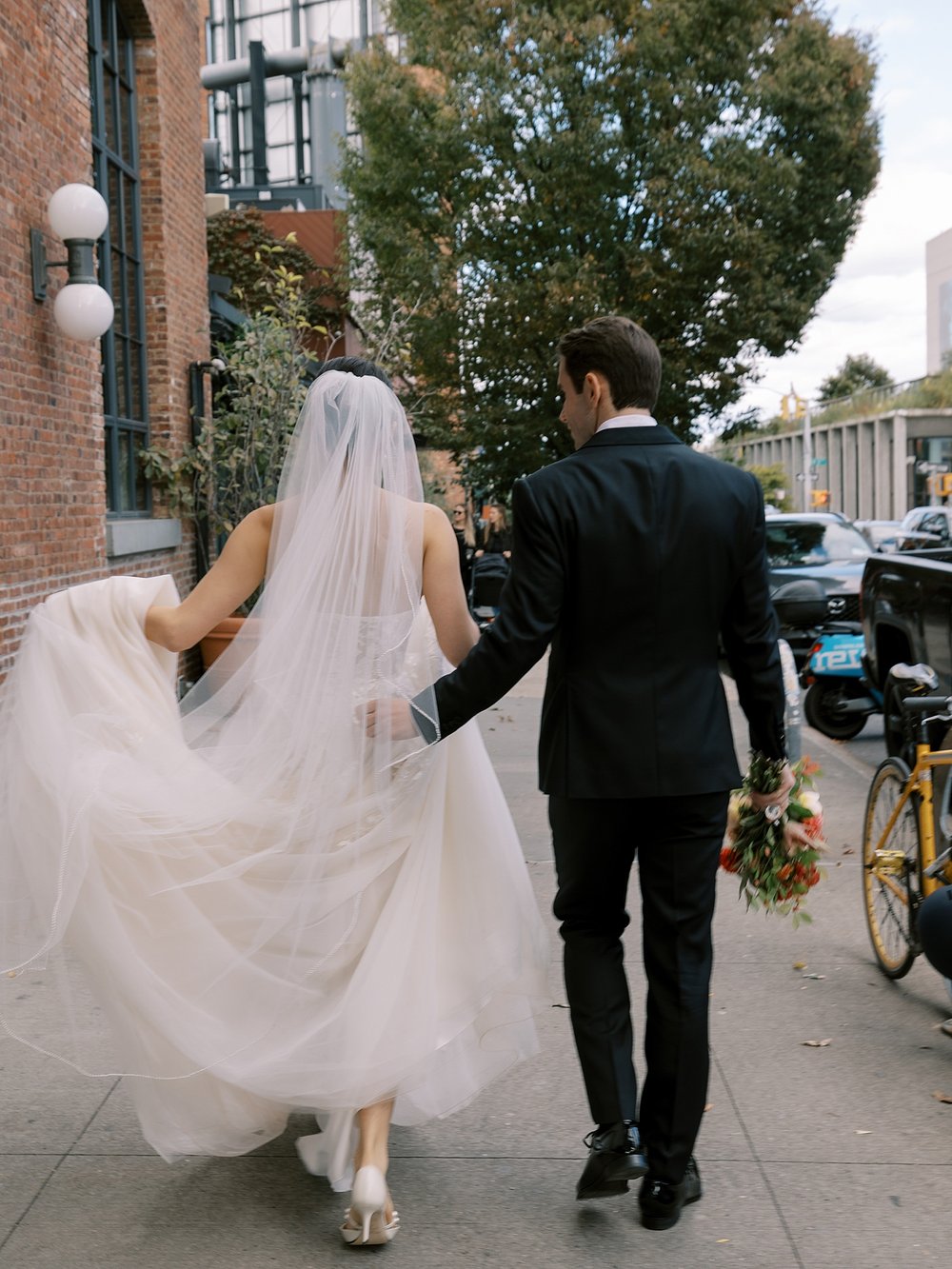 groom helps bride lift skirt to walk on NYC sidewalk outside hotel