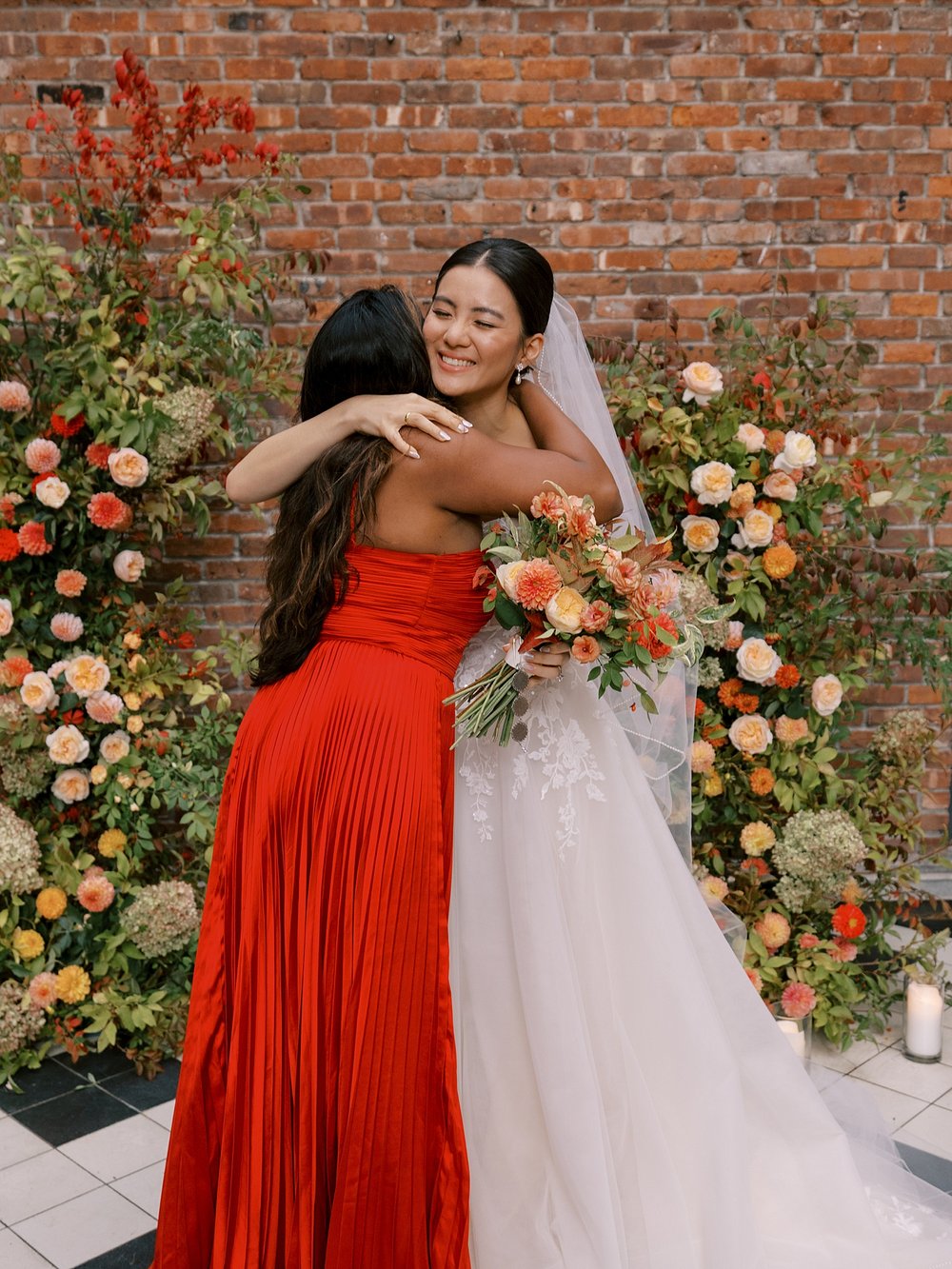 bride hugs guest in red dress in front of brick wall at the Wythe Hotel