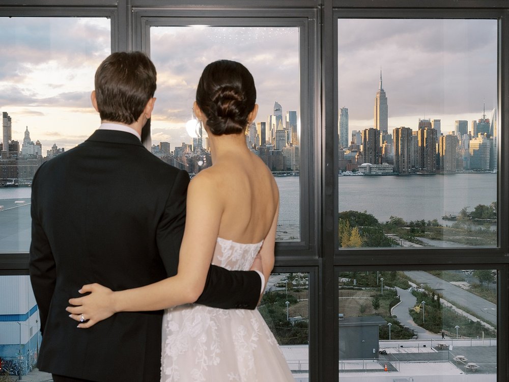 newlyweds hug in front of large windows overlooking Brooklyn