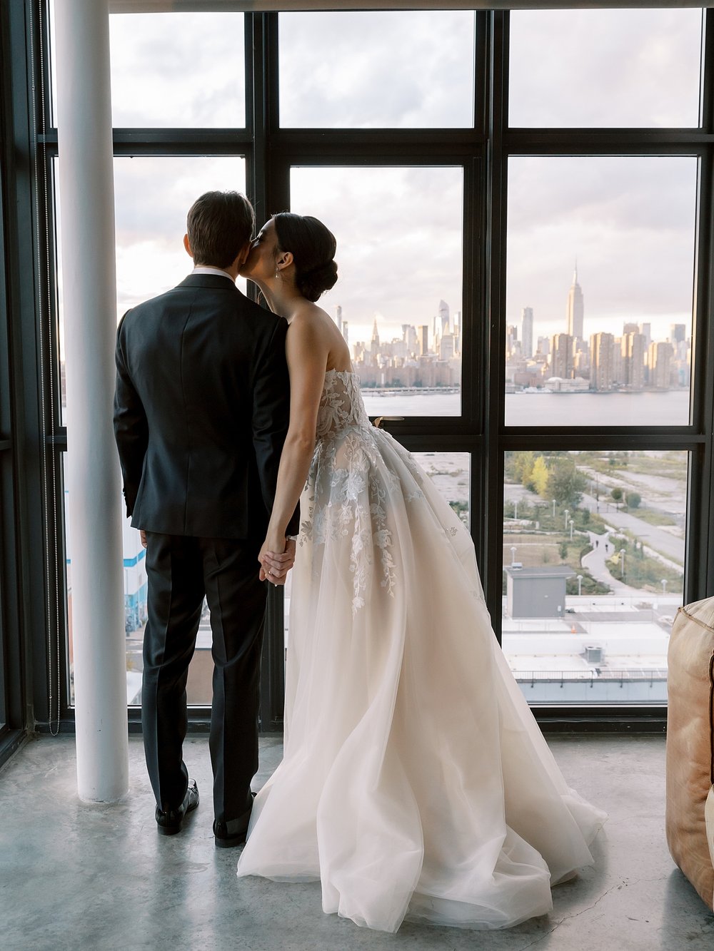 bride leans to kiss groom's cheek in front of large windows overlooking Brooklyn