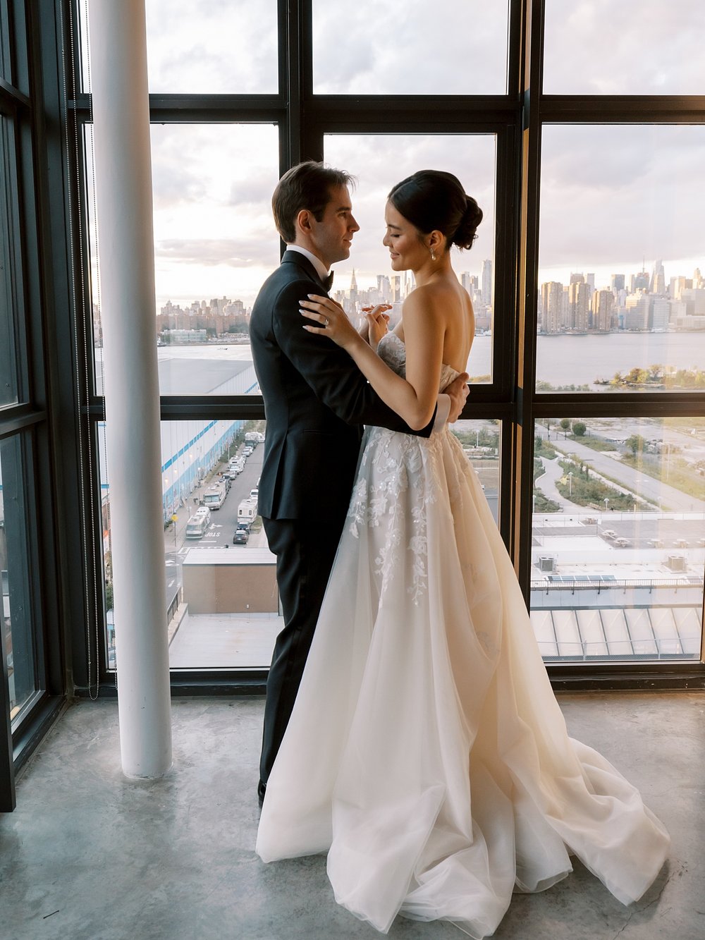 bride and groom hug facing each other in front of large windows overlooking Brooklyn