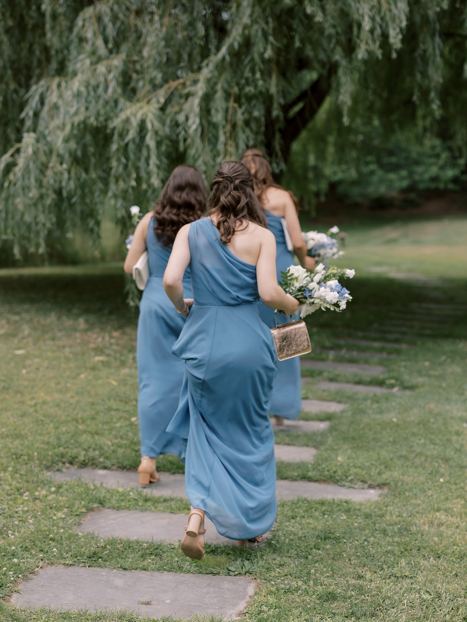bridesmaids in blue gowns walk by weeping willow 