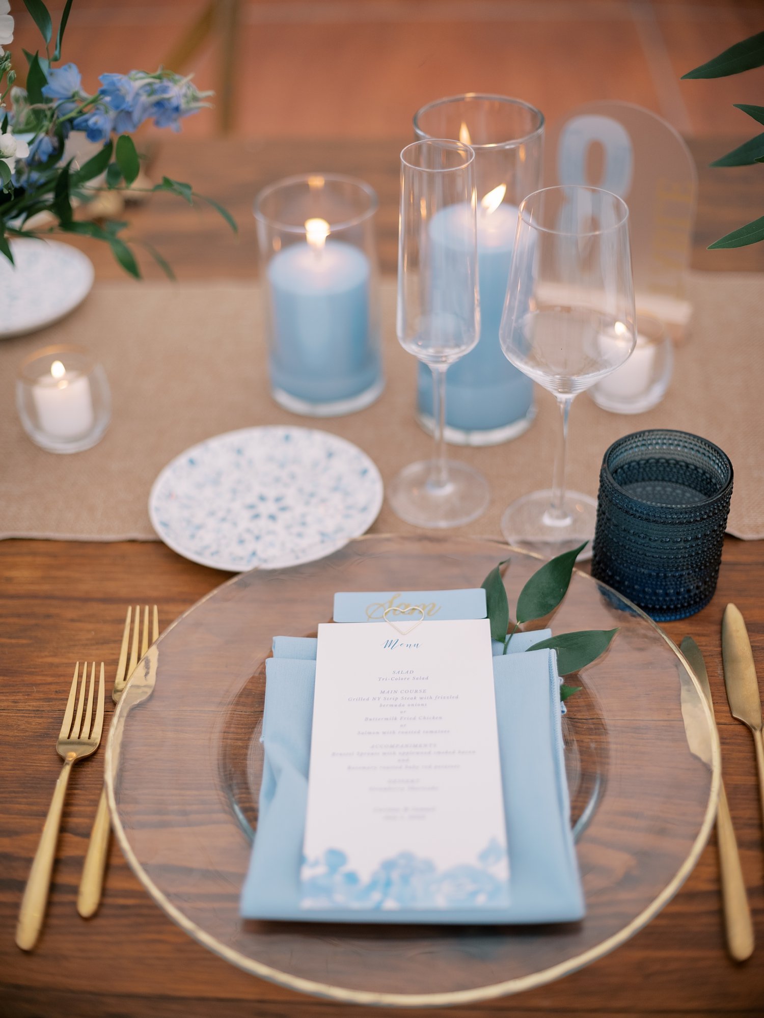 place setting on wooden table with clear plate with gold rim and blue menu card