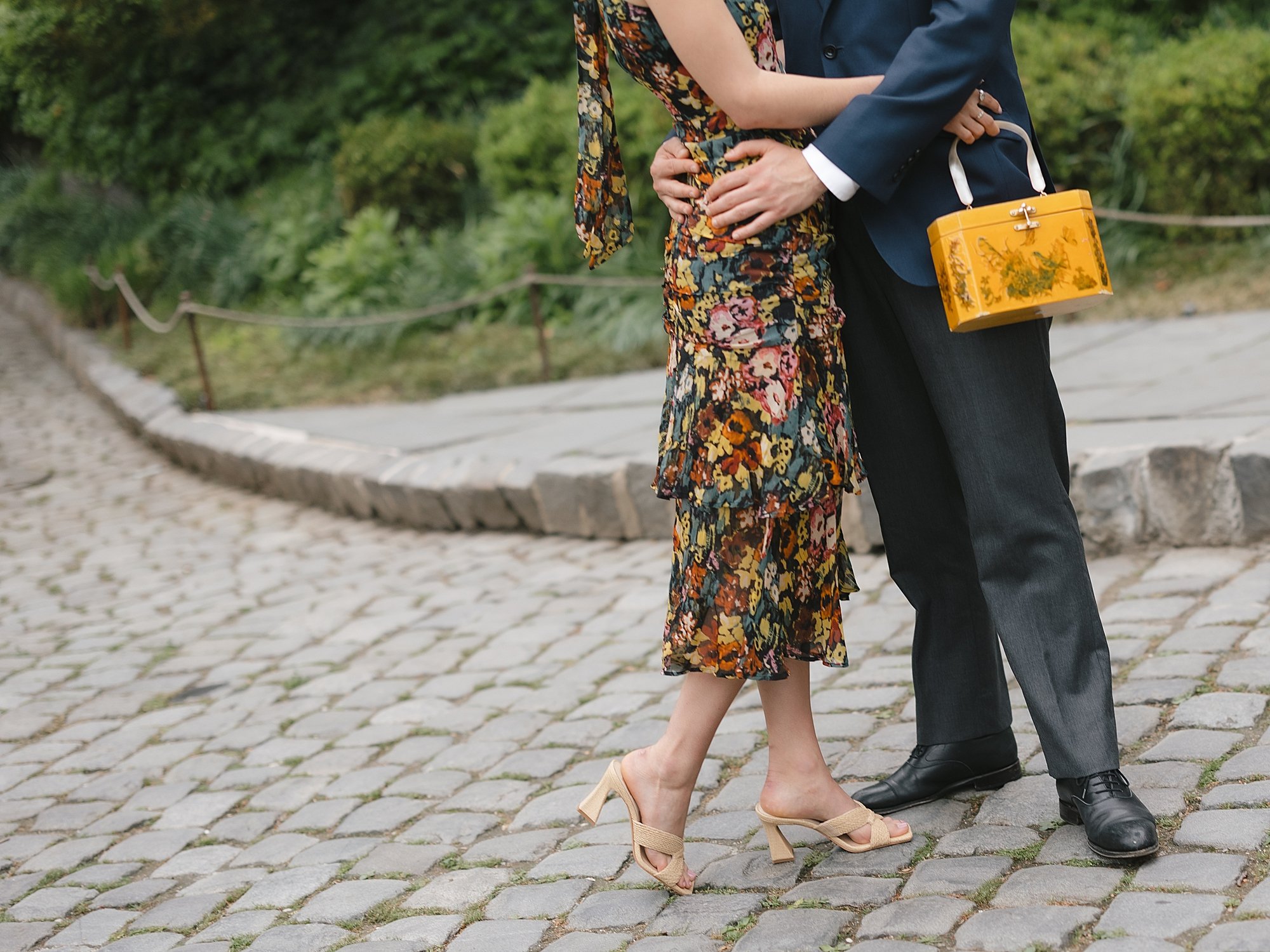 man holds woman in floral dress around the waist on hill inside Fort Tryon Park