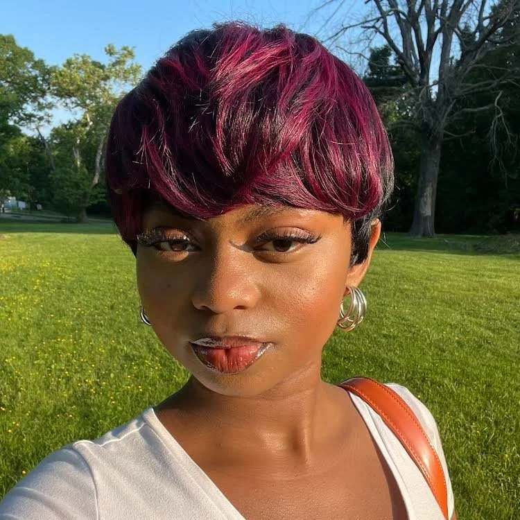 A woman with short hair dyed pink and black, standing outdoors in a park on a sunny day, wearing silver hoop earrings and a white top with a brown strap over her shoulder.