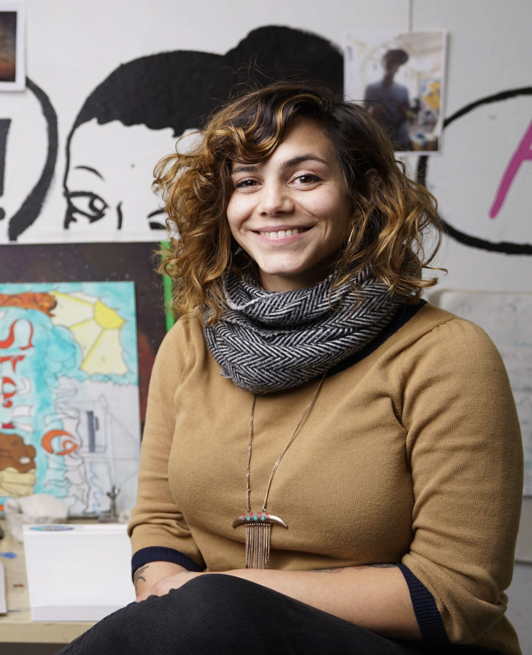 A woman with curly brown hair smiling at the camera, wearing a tan sweater, a patterned black and white scarf, and a necklace, in an art studio with colorful paintings in the background.
