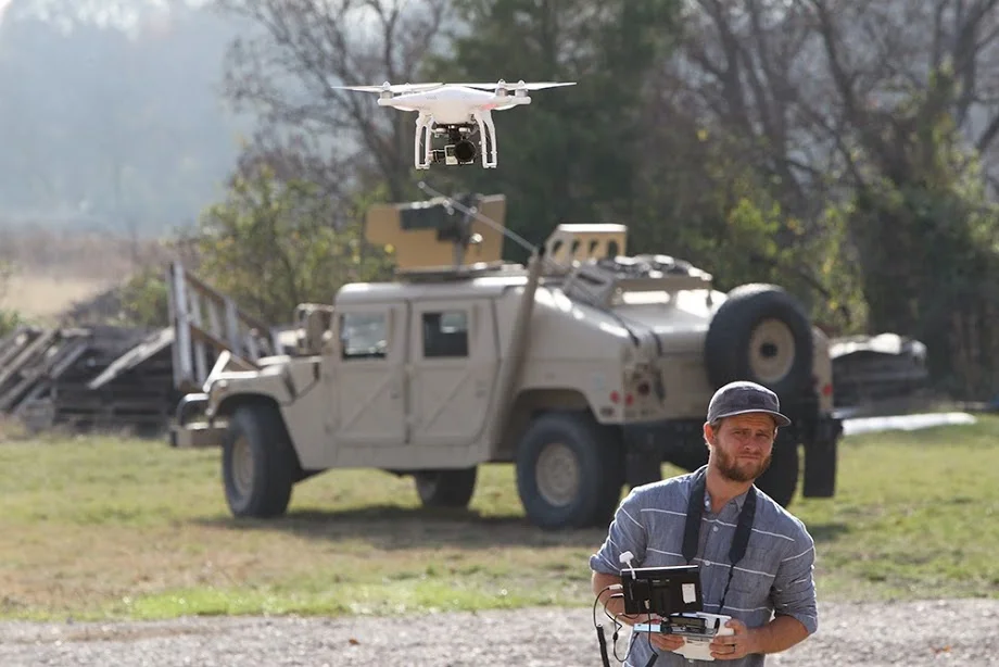 A man operating a drone with a remote control in an outdoor setting, with an off-road vehicle in the background.
