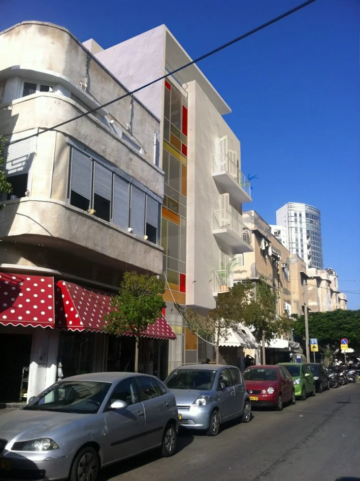 MARKET PLACE We are standing on Rambam street, two building away to the "Carmel" market, overlooking this new project's site. Overlooking the project and smelling the market. Seeing the dealers stands in the nearby pedestrian street, but stealing glances at the market. The market was there first. And probably will remain long after the next municipal project. When arriving back at the office, we spread our sketch books, and it was clear to us that it is not possible to design another typical Tel Avivian building in the front row with the well known market. A little wood coating and some popping windows just won't do it. We designed on the ground floor a store out of consideration of the existing urban façade, and a joyful colorful glass along the street façade. In another market, the one in Jaffa, we found a man that can still produce old fashion shutters. Metal doors with small cracks for air flow and natural light penetration. We told our selves, that one day, when we are older and the kids will be all grown up, we will buy an apartment by the market and we will go strolling the market on early Friday mornings just before all the crowd arrives. And we will feel 24 years old. Again. Site area: 325 sq | built area: 890 sq Architect in charge: Galit Shacham | Entrepreneur: "Kennedy Realestate" | Illustrations: Anderman Architects