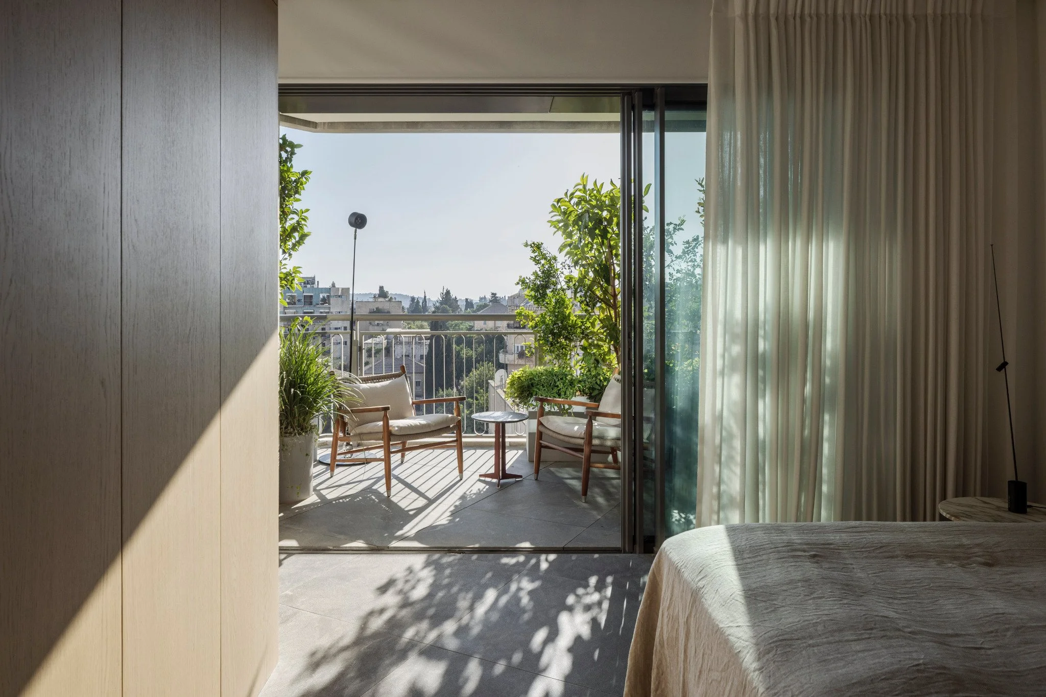 A balcony with outdoor furniture, lush green plants, and a city view with apartment buildings, as seen from inside a bedroom with curtains and a bed.