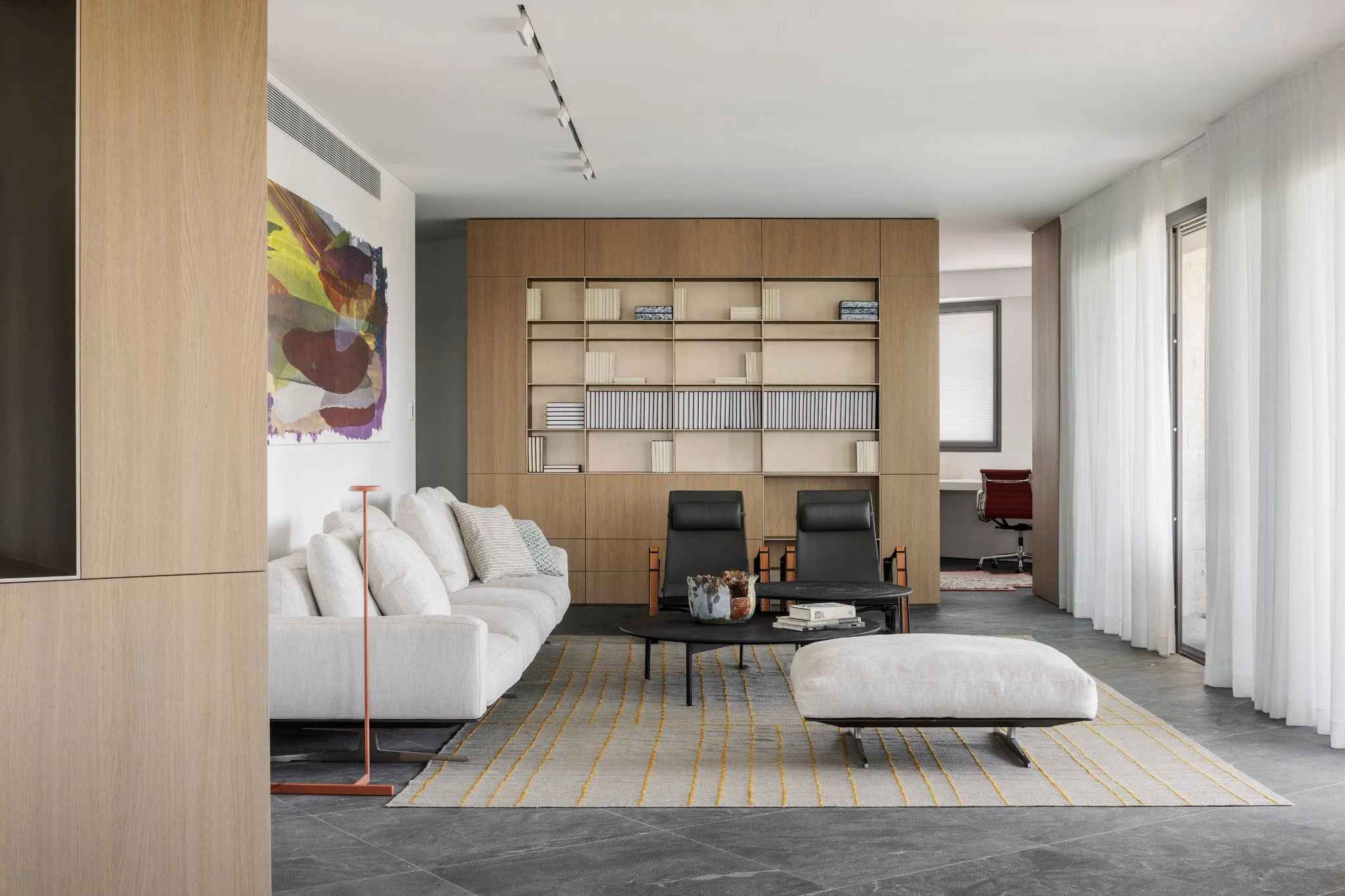 Modern living room with a white sofa, black chairs, a coffee table, wooden bookshelf, abstract art, and large windows with sheer curtains.