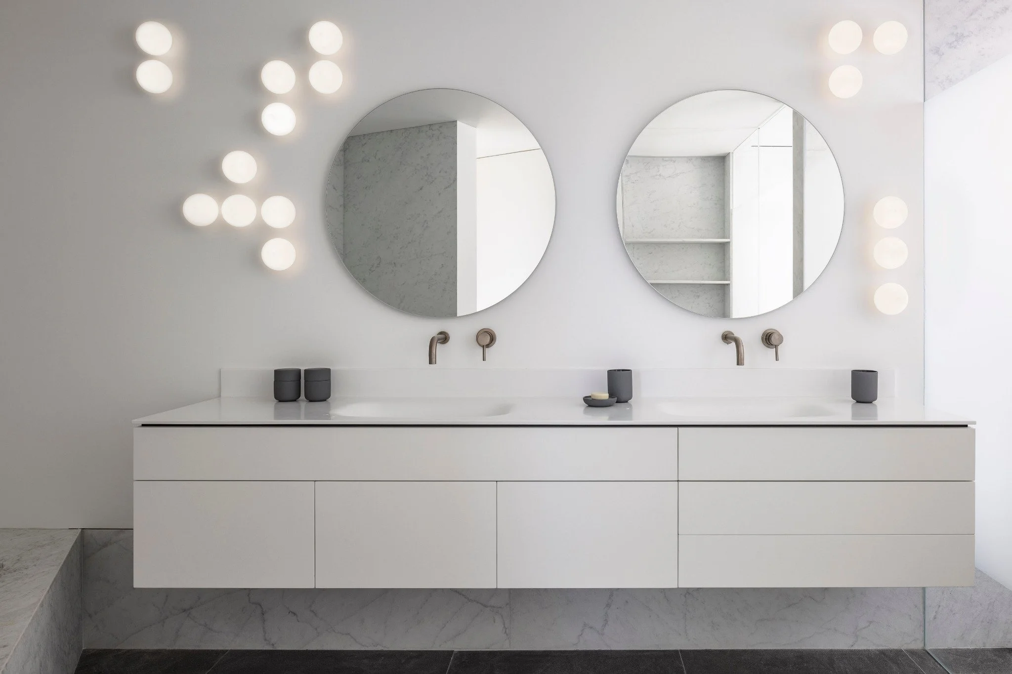 Minimalist bathroom with two round mirrors, white vanity, black containers, and wall-mounted faucets, illuminated by round wall lights.