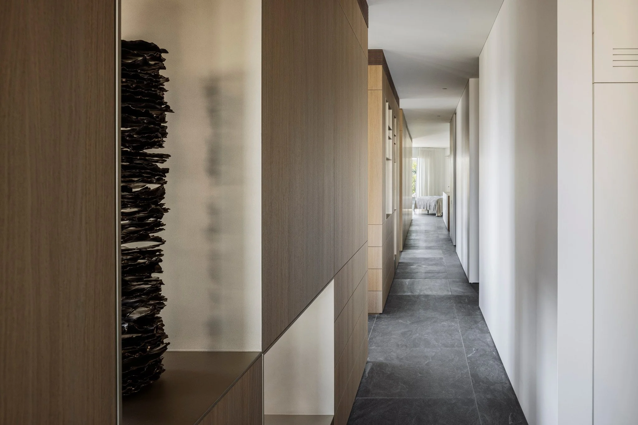 A modern hallway with wooden wall panels on the left, white walls on the right, and a small decorative sculpture resembling a vertical stack of dark leaves or feathers on the left wall. The hallway leads to a bright room with large windows and a bed.