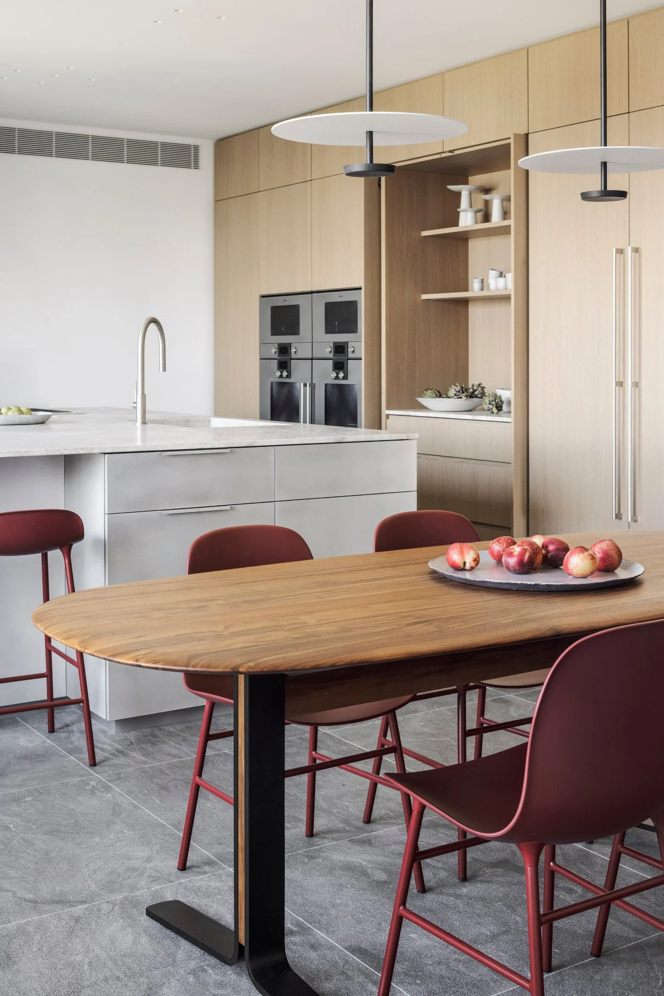 Modern kitchen with wooden cabinets, white countertop, built-in ovens, and a dining table with red chairs. Apples are on the table, and decorative dishes are on the open shelves.