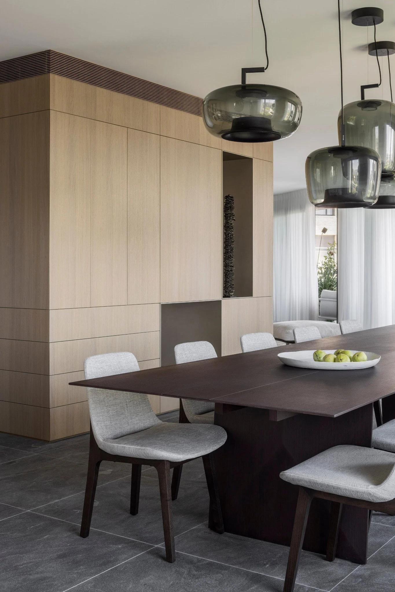 Modern dining room with a dark wood table surrounded by gray upholstered chairs, a white bowl with green apples on the table, a large wooden wall with built-in shelving, and pendant lights hanging from the ceiling, with a window and white curtains in